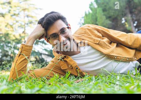 Smiling bearded ethnic man in eyeglasses lying on green grass with yellow leaves and looking at camera Stock Photo