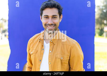 Smiling young ethnic bearded man in yellow shirt standing near blue wall in park and looking at camera Stock Photo