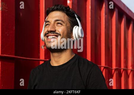Cheerful man with mustache and in black outfit while listening to favorite song on headphones against red metal fence looking away Stock Photo