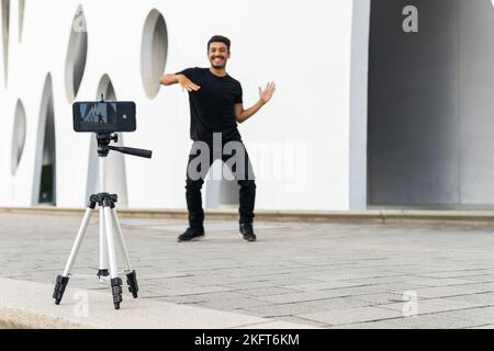 Full body of happy male blogger dancing on street while recording video on smartphone fixed on tripod Stock Photo