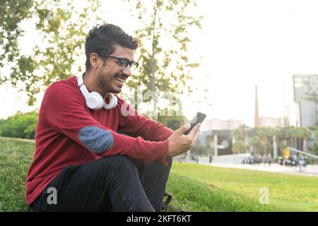 Smiling young concentrated male with headphones on neck sitting on grass in park browsing on mobile phone Stock Photo