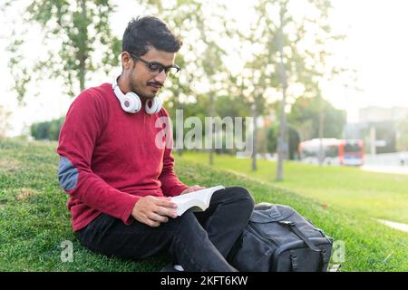 Ground level of young concentrated male with headphones on neck sitting on grass in park reading novel Stock Photo
