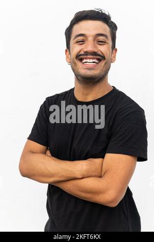 Positive bearded man with mustache standing on isolated background with hands crossed in black t shirt smiling and looking at camera Stock Photo