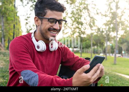 Smiling young concentrated male with headphones on neck sitting on grass in park browsing on mobile phone Stock Photo