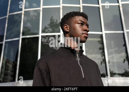 Ethnic young guy in black clothes standing on street near glass building wall and looking away thoughtfully Stock Photo