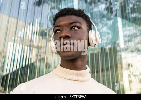 Low angle of African American male in casual clothes listening to music via wireless headphones against glass building Stock Photo