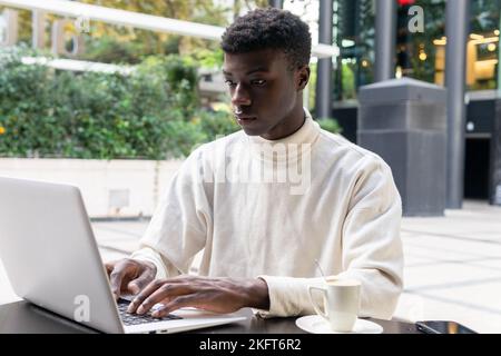 Concentrated African American male freelancer in casual clothes sitting at table with cup of coffee and typing on laptop at outdoor cafe Stock Photo