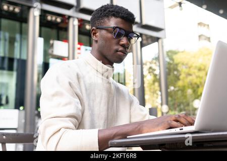 Concentrated African American male freelancer in casual clothes sitting at table with cup of coffee and typing on laptop at outdoor cafe Stock Photo