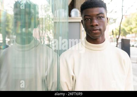 Portrait of serious African American male in casual clothes looking at camera while leaning on glass wall Stock Photo