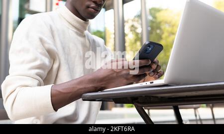 From below of handsome African American male in glasses and sweater texting on mobile phone while sitting with laptop at table against modern building Stock Photo
