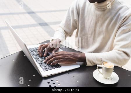 Crop African American male freelancer in casual clothes sitting at table with cup of coffee and typing on laptop at outdoor cafe Stock Photo