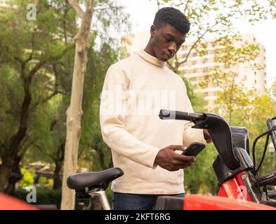 Low angle of African American male in casual clothes renting electric bicycle using mobile phone app in city Stock Photo