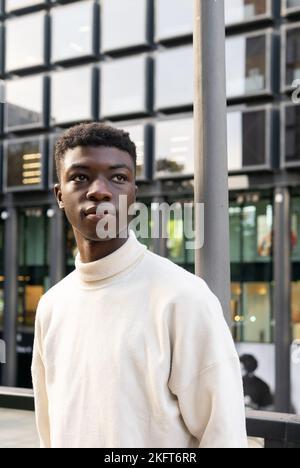 Young African American guy in light clothes standing against building in city and looking away Stock Photo