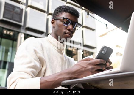 From below of handsome African American male in glasses and sweater texting on mobile phone while sitting with laptop at table against modern building Stock Photo