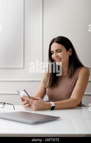 Positive adult female entrepreneur with long dark hair in casual clothes and smart watch smiling while sitting at desk with laptop and eyeglasses and Stock Photo