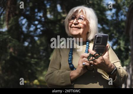 Smiling elderly lady in glasses and beads browsing on mobile phone standing near green trees while looking away Stock Photo
