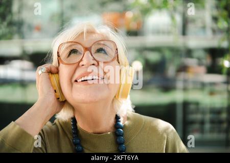 Smiling elderly lady in eyeglasses with white hair standing on street and listening to music via yellow wireless headphones while looking away Stock Photo