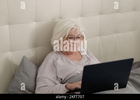 Happy elderly lady in eyeglasses lying on soft bed with gray bedclothes and typing information on laptop at home Stock Photo