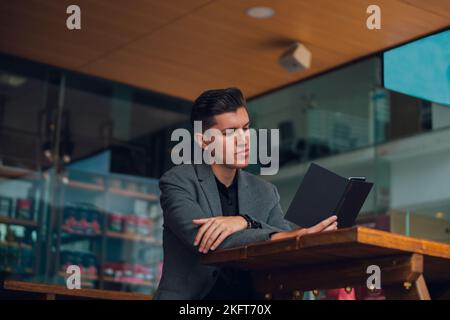 Concentrated young man in formal outfit sitting at table in cafeteria while reading notes in planner Stock Photo