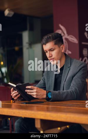Concentrated young man in formal outfit sitting at table in cafeteria while reading notes in planner Stock Photo