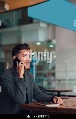 Smiling young man having phone call while resting at table with notebook communicating spending time in cafe Stock Photo
