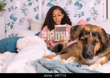 Teenage hispanic female in pajamas using a tablet computer while sitting on bed at home with her dog pet Stock Photo