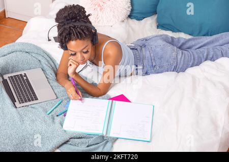 Top view of teenage hispanic female in casual clothes and headphones writing down on notebook while lying on bed at home next to laptop computer Stock Photo