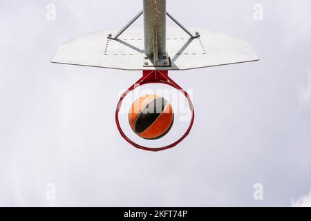 From below of orange basketball falling into round hoop with net placed against cloudy sky in city Stock Photo