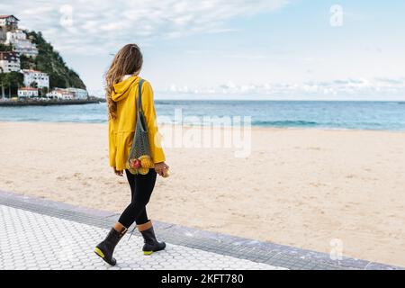 Back view of anonymous female in bright yellow coat watching waves of sea washing sandy beach in Donostia of Spain Stock Photo