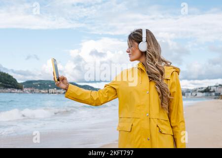 Young female in yellow raincoat having video call on mobile phone on foamy sea of Spain Stock Photo