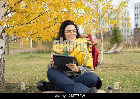 Happy young female wrapped in blanket sitting on bench and browsing tablet while sitting in autumn park Stock Photo