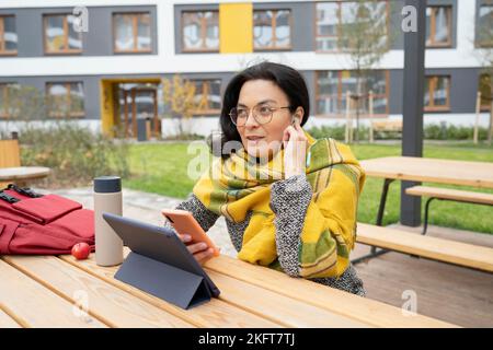 Young female using earphones and mobile phone while sitting at table with tablet and lunch snack in campus park in fall season Stock Photo