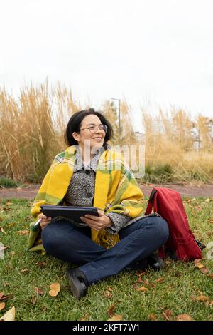 Full body adult female designer in warm jacket and plaid on shoulders sitting on grassy lawn and looking around for inspiration in fall season in natu Stock Photo