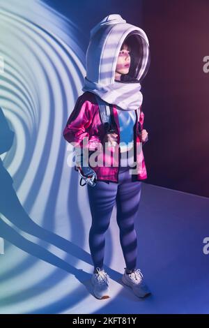 Young Asian female with backpack wearing cosmonaut helmet looking away in calm solitude standing in pink glowing light on foil background Stock Photo