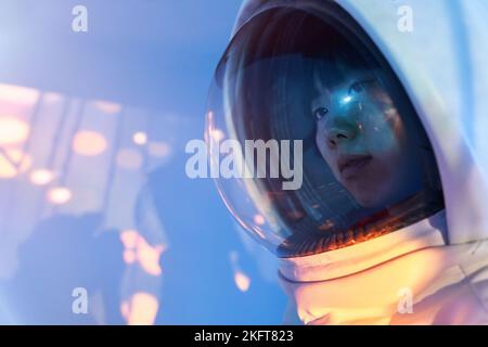 Young Asian female wearing cosmonaut helmet looking away in calm solitude standing in pink glowing light on foil background Stock Photo