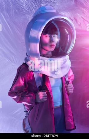 Young Asian female with backpack wearing cosmonaut helmet and closing eyes in calm solitude standing in pink glowing light on foil background Stock Photo