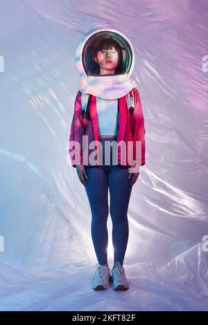 Young Asian female with backpack wearing cosmonaut helmet and closing eyes in calm solitude standing in pink glowing light on foil background Stock Photo