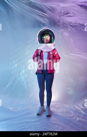 Young Asian female with backpack wearing cosmonaut helmet looking away in calm solitude standing in pink glowing light on foil background Stock Photo