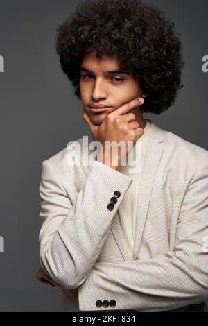 Pensive young Hispanic man in stylish suit with curly hair touching chin and looking at camera while pondering against gray background Stock Photo