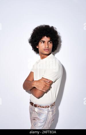 Young Hispanic man in white t shirt and silver pants with curly hair crossing arms and looking away over shoulder against gray background Stock Photo