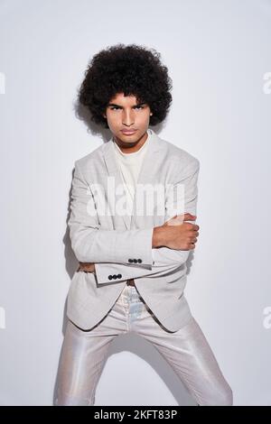 Young Hispanic male in stylish clothes with curly hair with arms crossed and laughing against gray background Stock Photo
