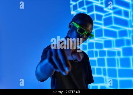 Self assured young African American male pointing and looking at camera in ultraviolet light with 3D cubes Stock Photo