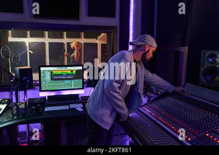 Side view of focused male recording engineer standing at workplace with computer while musician singing song on microphone Stock Photo