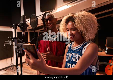 African American woman with Afro hairstyle using smartphone near microphone and black male singing song Stock Photo