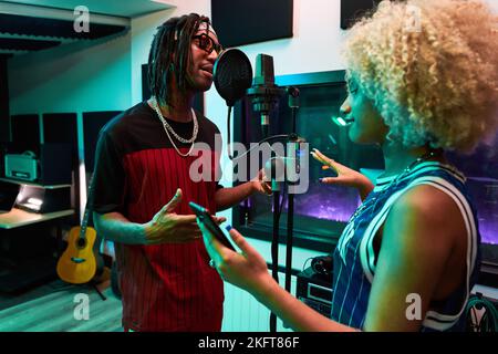 Side view of African American woman with Afro hairstyle using smartphone near microphone and black male singing song Stock Photo
