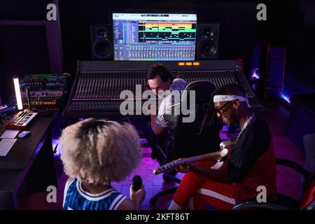 From above of multiracial people gathering in recording studio with glowing neon lights while black man playing electric guitar and female singing on Stock Photo