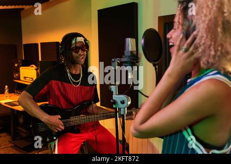 Male African American musician in headphones playing guitar while female partner recording song on microphone Stock Photo