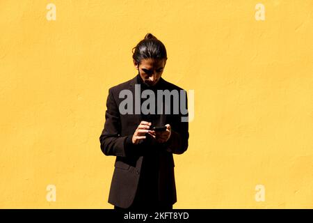 Hispanic male in black suit browsing social media on cellphone against bright yellow wall Stock Photo