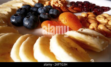 Fresh and dried fruits and nut dish, banana sliced, blueberries and apricot in the center and of the white plate with nut. Stock Photo