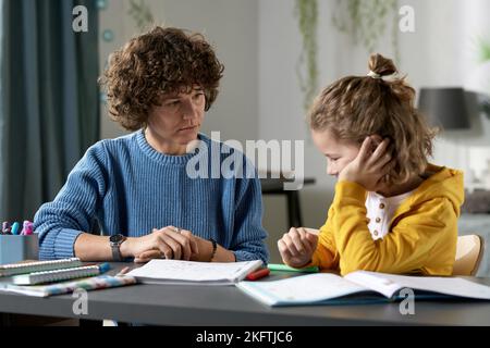 Young teacher teaching difficult child at home, they sitting together at desk with books and talking Stock Photo